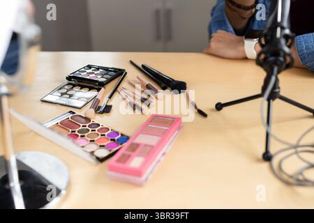 Influenceur non binaire arrangeant des palettes de fard à paupières et de blush avec des pinceaux près du miroir, micro sur le bureau Banque D'Images