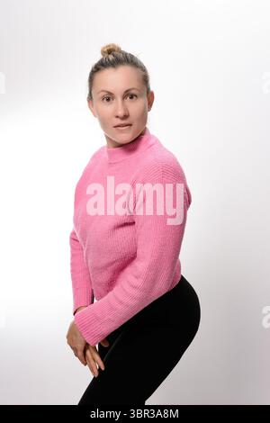 Une femme avec ses cheveux dans un chignon, portant un pull rose et des leggings noirs, se tient sur un fond blanc. Elle est légèrement penchée en avant, créant un d. Banque D'Images
