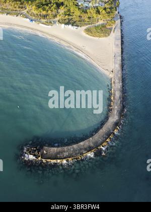 Vue aérienne de la jetée courbée qui s'étend dans l'océan turquoise depuis la plage de sable, une escapade tranquille sous le soleil chaud, Bal Harbour, Floride, États-Unis. Banque D'Images