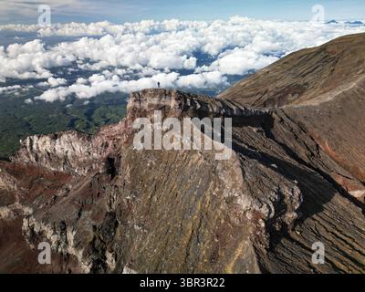 Vue aérienne d'une figure solitaire debout au sommet d'un précipice rocheux déchiqueté, surplombant les vallées verdoyantes et les nuages lointains et moelleux, volcan Agung, Karangasem, Bali, Indonésie. Banque D'Images