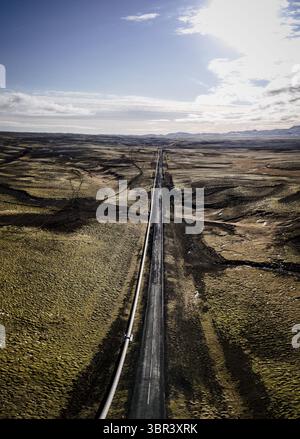 Vue aérienne d'une route droite coupe le paysage accidenté, contrastant l'asphalte sombre avec des champs dorés, sous un vaste ciel, Mosfellsbaer, Höfuðborgarsvæðið, Islande. Banque D'Images