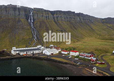 Vue aérienne des cascades descendant les falaises accidentées, embrassant le village pittoresque niché en contrebas dans un mélange harmonieux de nature et de présence humaine, Westfjords, Islande. Banque D'Images