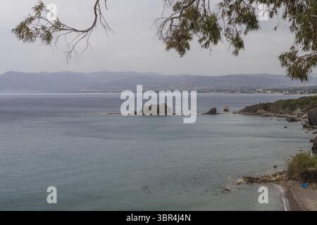 Vue sur les eaux turquoises tranquilles rencontrer la côte accidentée, encadrée par de délicates branches d'arbres sur un fond de montagnes brumeuses, Paphos, Paphos, Chypre. Banque D'Images