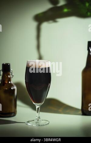Bière sombre déversant d'une bouteille à un verre avec tête en mousse sur fond vert coloré, ombres vives des feuilles de palmier Banque D'Images