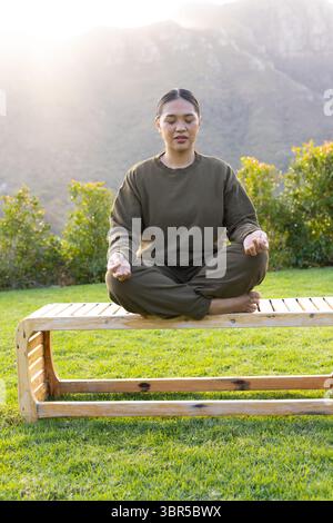 Femme asiatique méditant pieds nus croisés sur un banc en bois dans une pelouse herbeuse avec des haies vertes Banque D'Images