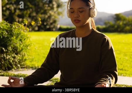 Femme asiatique adulte moyenne portant un sweat-shirt olive méditant sur le rebord en pierre par pelouse portant des écouteurs Banque D'Images