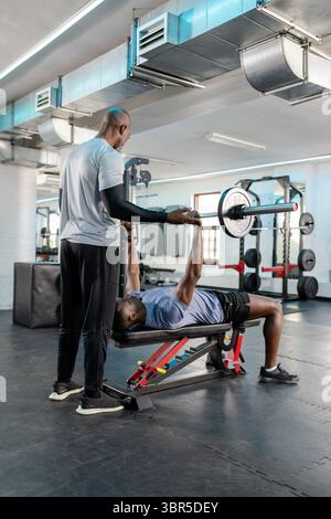 Entraîneur de fitness afro-américain repérant le client faisant la presse de banc avec barre sur banc à la salle de gym Banque D'Images