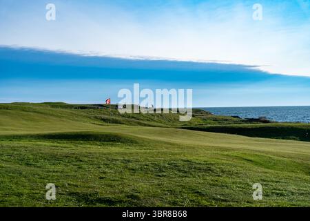 Vue sur les collines verdoyantes du parcours de golf rencontre le vaste océan bleu sous un ciel lumineux, Vestmannaeyjabær, Vestmannaeyjabær, Islande. Banque D'Images