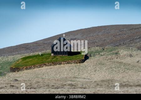 Vue d'une église sombre et en bois, Krýsuvíkurkirkja, avec des croix blanches, se dresse au sommet d'un boulevard herbeux, entouré d'un mur de pierre bas dans un vent désolé Banque D'Images
