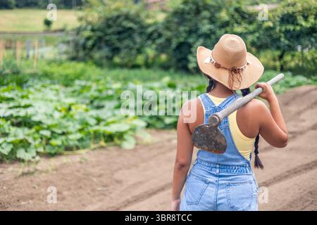 Une jeune agricultrice latino-américaine porte une houe sur son épaule tout en marchant dans un champ cultivé, prête à travailler la terre et à cultiver Banque D'Images