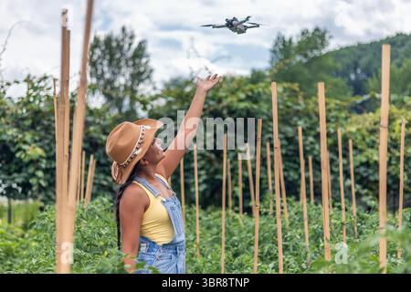 Jeune agricultrice latino-américaine portant un chapeau de paille et une combinaison en denim lançant un drone au-dessus de ses plants de tomates dans un champ, en utilisant des technologies modernes Banque D'Images
