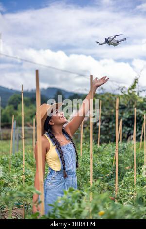 Verticale jeune agricultrice latino-américaine avec chapeau de paille et salopettes en denim contrôlant un drone volant au-dessus des plants de tomates dans sa ferme, en utilisant moderne Banque D'Images