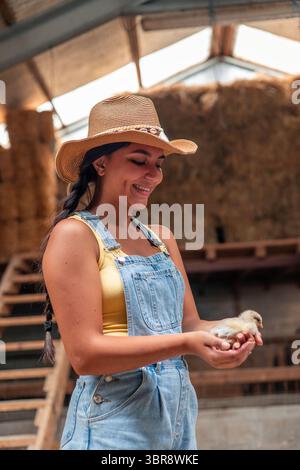 Souriante jeune agricultrice latine portant une combinaison en denim et un chapeau de paille, tenant doucement un petit poussin dans ses mains tout en étant entouré par l'ambiance chaleureuse Banque D'Images