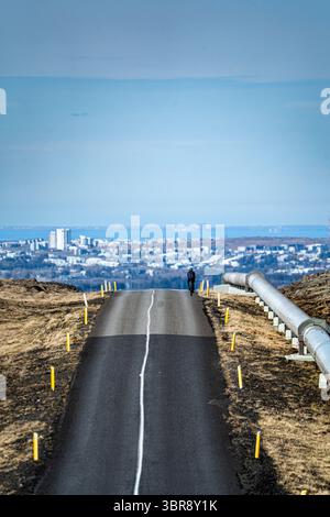 Vue d'une route noire dure coupant à travers des champs dorés vers la ville lointaine sous un ciel pâle, avec un grand pipeline sur le côté, Nesjavellir, Gr Banque D'Images
