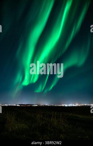 Vue des danses vertes éclatantes des aurores boréales à travers le ciel nocturne, projetant une lueur éthérée sur le paysage sombre, Reykjavík, Reykjavíkurborg, Islande. Banque D'Images