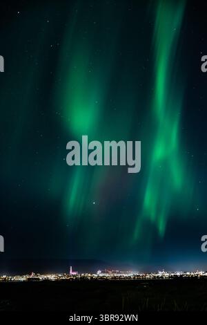 Vue aérienne de l'envoûtante aurore boréale danse à travers le ciel nocturne au-dessus des lumières lointaines de la ville, une symphonie de teintes vertes et sombres, Islande. Banque D'Images