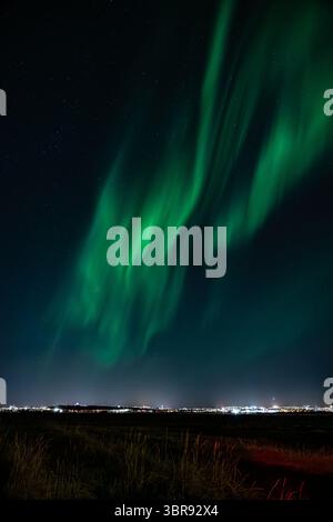 Vue aérienne d'une aurore boréale vibrante danse à travers le ciel sombre, projetant une lueur verte éthérée sur les lumières lointaines de la ville, Islande. Banque D'Images