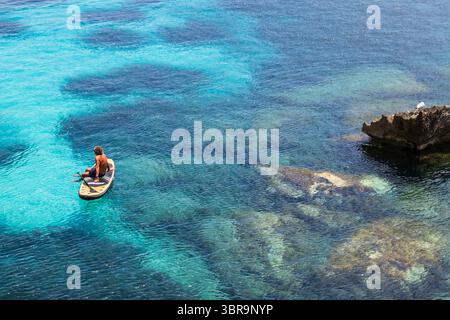Homme assis sur une planche de surf dans une mer turquoise cristalline avec des fonds marins visibles, des rochers et des herbiers marins Banque D'Images