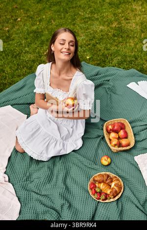 Une belle jeune femme est assise sur une couverture de pique-nique, savourant une pomme entourée de collations. Banque D'Images