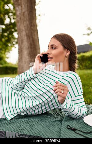 Une jeune femme se détend sur une couverture de pique-nique, profitant d'un appel dans le plein air ensoleillé. Banque D'Images
