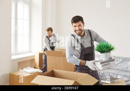Équipe de déménageurs heureux, groupe de service de deux hommes souriants tenant et portant des boîtes en carton, souriant Banque D'Images