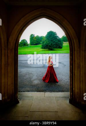 Une femme dans une robe rouge marche à travers une arche, avec un paysage vert en arrière-plan. Banque D'Images