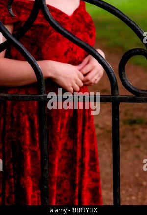 Une femme dans une robe rouge se tient derrière une porte en fer forgé noir, ses mains serrées ensemble. Banque D'Images