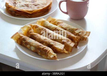 Crêpes roulées avec garniture de fromage cottage servies sur assiette blanche. Mug rose en arrière-plan soft focus. Petit déjeuner confortable ou idée de dessert léger Banque D'Images