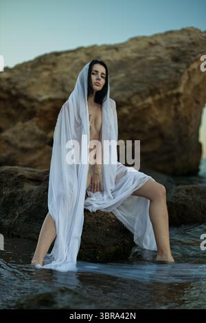 Serene moment capture une figure brune, enveloppée dans un tissu blanc fluide, assis sur un rocher au bord de l'océan le soleil se couche sur l'horizon, illuminant la scène paisible Banque D'Images