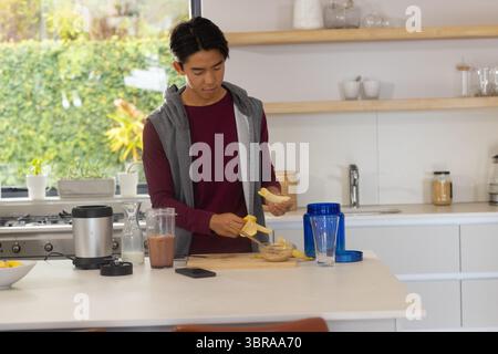 Homme asiatique pelant la banane sur le bol de beurre d'arachide à l'îlot de cuisine avec mélangeur, poudre de protéine Banque D'Images