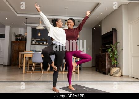 Diverses amies féminines tenant l'arbre posent sur des tapis de yoga dans le salon et la salle à manger ouverts Banque D'Images