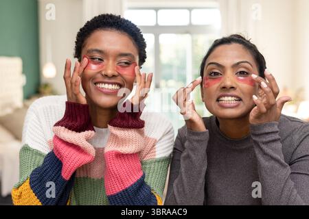 Diverses amies féminines appliquant des patchs sous les yeux de gel rouge sur le canapé dans le salon avec fenêtre Banque D'Images