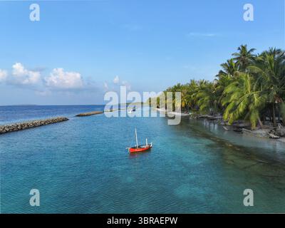 Vue aérienne d'un voilier rouge solitaire flottant sur les eaux turquoises près de la côte bordée de palmiers de Kendhoo, atoll de Baa, Maldives. Banque D'Images