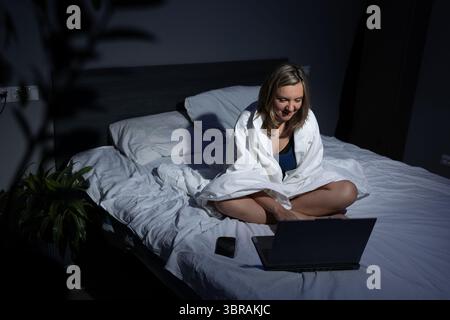 Une femme est assise les jambes croisées sur son lit la nuit, enveloppée dans une couverture tout en travaillant sur son ordinateur portable. La douce lueur de l'écran illumine son expression focalisée et la pièce confortable. Banque D'Images