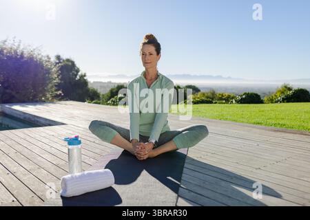 Femme senior exécutant l'étirement papillon assis sur le tapis de yoga de terrasse arrière portant des vêtements de sport verts Banque D'Images