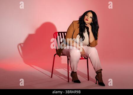 Femme assise sur une chaise en métal rouge dans le studio reposant menton sur la main tout en portant un blazer camel-brun Banque D'Images