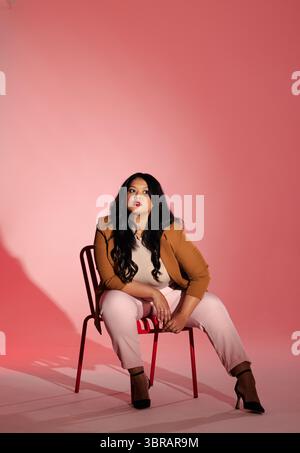 Femme asiatique posant sur une chaise en métal rouge dans un studio de photographie avec fond rose et ombres nettes Banque D'Images