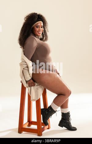 Femme afro-américaine assise sur le côté sur le tabouret dans le studio portant un body taupe et des bottines Banque D'Images