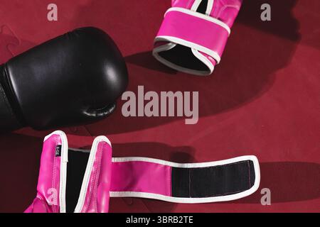 Gant de boxe noir couché sur le plancher de l'anneau rembourré rouge avec deux gants roses exposant la sangle détachée Banque D'Images