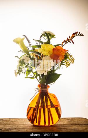 Un bouquet coloré mettant en vedette des lis calla, des roses et d'autres fleurs mélangées dans un vase ambre unique, placé sur du bois rustique sur un fond blanc propre. Banque D'Images