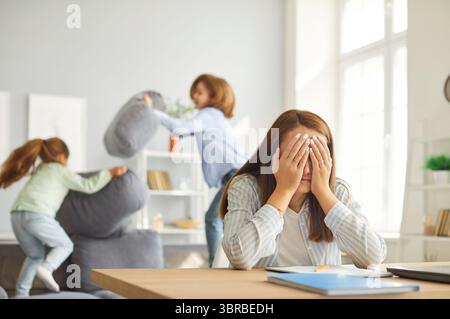 Mère épuisée assise au bureau avec la tête dans les mains alors que les enfants sautent et jouent fort sur le canapé. Banque D'Images
