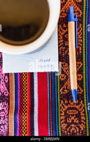Tasse à café avec une note disant bonjour sur une table colorée avec tissage Inca. Banque D'Images