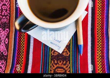 Tasse à café avec une note disant bonjour sur une table colorée avec tissage Inca. Banque D'Images