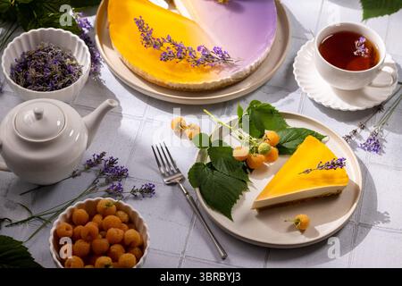 Tranche de cheesecake à la lavande servie dans une assiette avec des framboises jaunes, une tasse de thé, une théière et un bol de fleurs de lavande séchées Banque D'Images