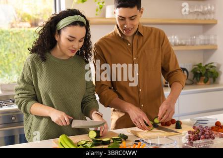 Couple diversifié hachant des légumes sur des planches à découper en bois à l'îlot de cuisine avec les couteaux du chef Banque D'Images