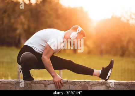 Un jeune homme en forme écoutant de la musique sur des écouteurs sans fil tout en faisant des exercices d'étirement avant de courir ou de s'entraîner dans le parc au coucher du soleil. Extérieur Banque D'Images