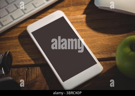 La conception plate montre un smartphone blanc reposant sur un bureau en bois avec clavier, souris, pomme verte Banque D'Images