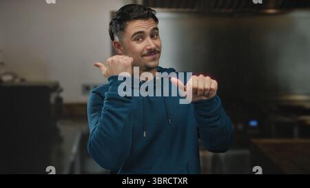 Jeune homme hispanique en sweat à capuche bleu souriant et pointant les pouces sur le côté dans le cadre de la cuisine offrant une atmosphère intérieure vibrante et un geste amical. Banque D'Images
