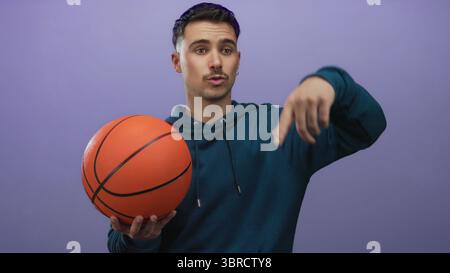 Jeune homme hispanique tenant le basket devant un mur isolé de fond violet, portant un sweat à capuche décontracté, pointant avec confiance, suggérant l'action ou moi Banque D'Images