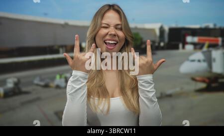 Femme blonde fait des gestes de cornes de roche avec les mains et les clins d'œil tout en souriant au terminal de l'aéroport ; rock fun metal énergie. Banque D'Images